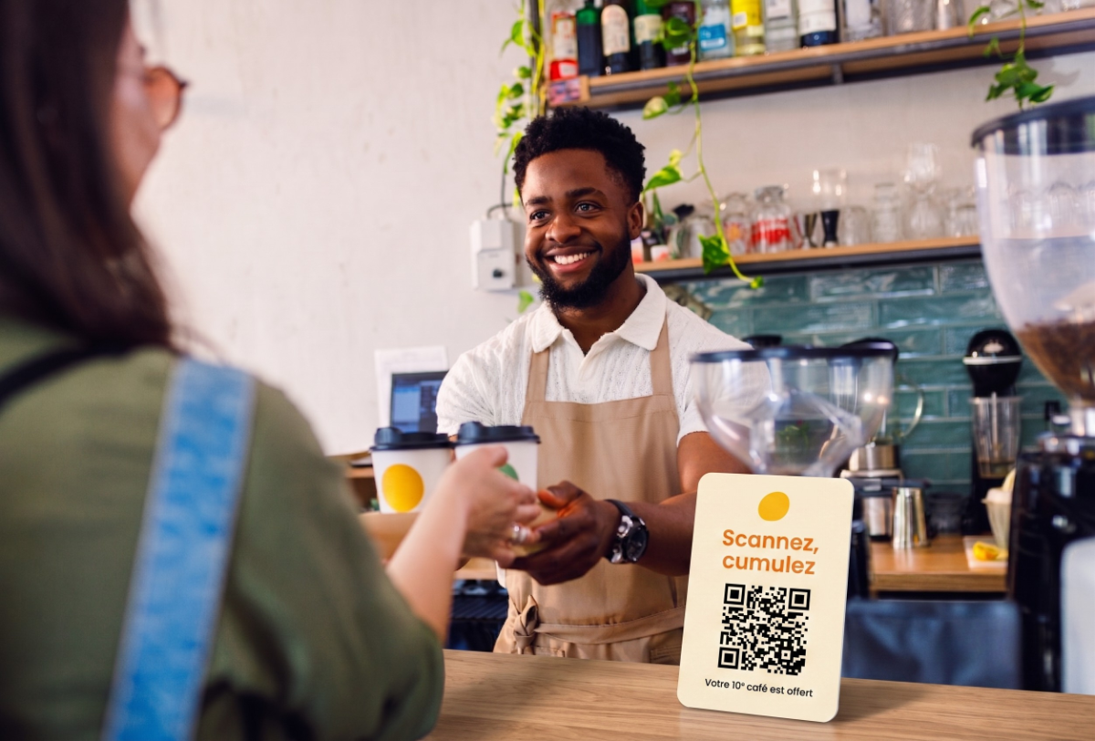 Un barista tend leur commande à une cliente, un chevalet Scannez, cumulez posé sur le comptoir